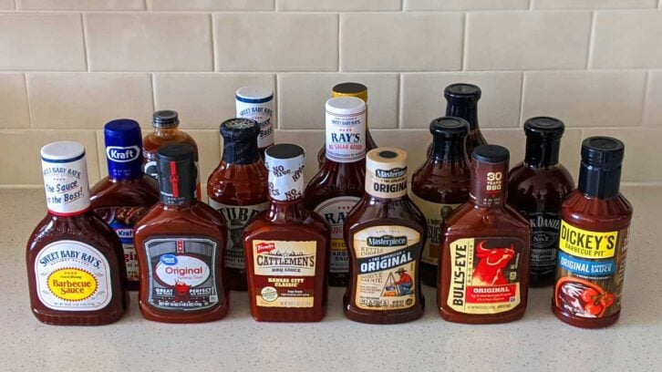 A variety of barbecue sauce bottles from different brands are arranged on a kitchen counter in front of a tiled backsplash.