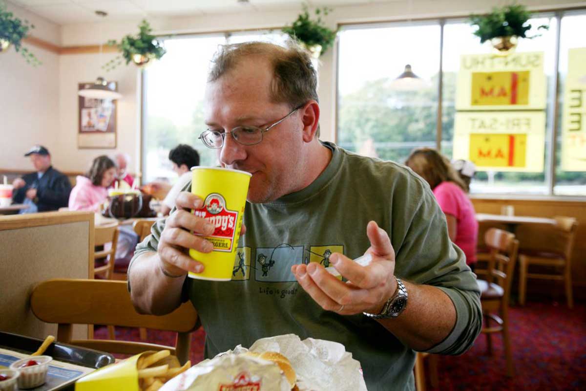 A man wearing glasses and a green shirt drinks from a large yellow Hardee’s cup—one of the most popular restaurant chains among boomers—while sitting at a table with food wrappers and a sandwich in front of him. Other diners are in the background.