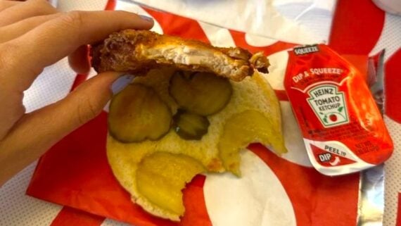 A hand holds a bitten fried chicken patty above a sandwich bun with pickle slices. Next to the sandwich, which sits on a red and white wrapper, is a Heinz ketchup packet—the scene could be from one of the worst chicken chains.