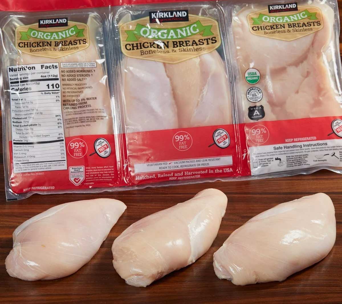 Three raw chicken breasts sit on a wooden surface in front of three packages of Kirkland Organic Boneless Skinless Chicken Breasts, a popular item often debated among lists of Costco products to avoid, with nutrition facts and labels clearly visible.