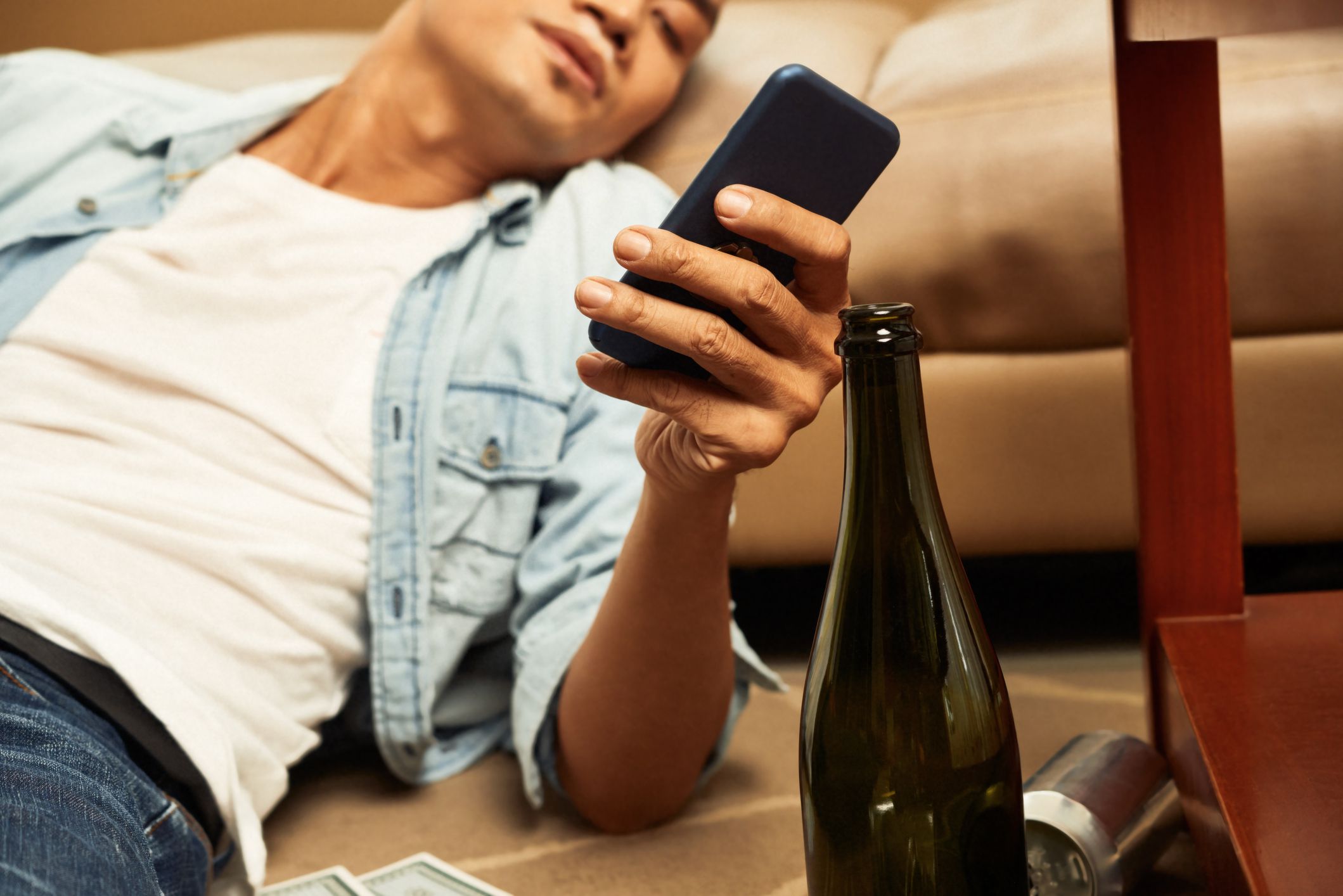 Addict young man lying on floor among alcohol bottles and can and texting friend