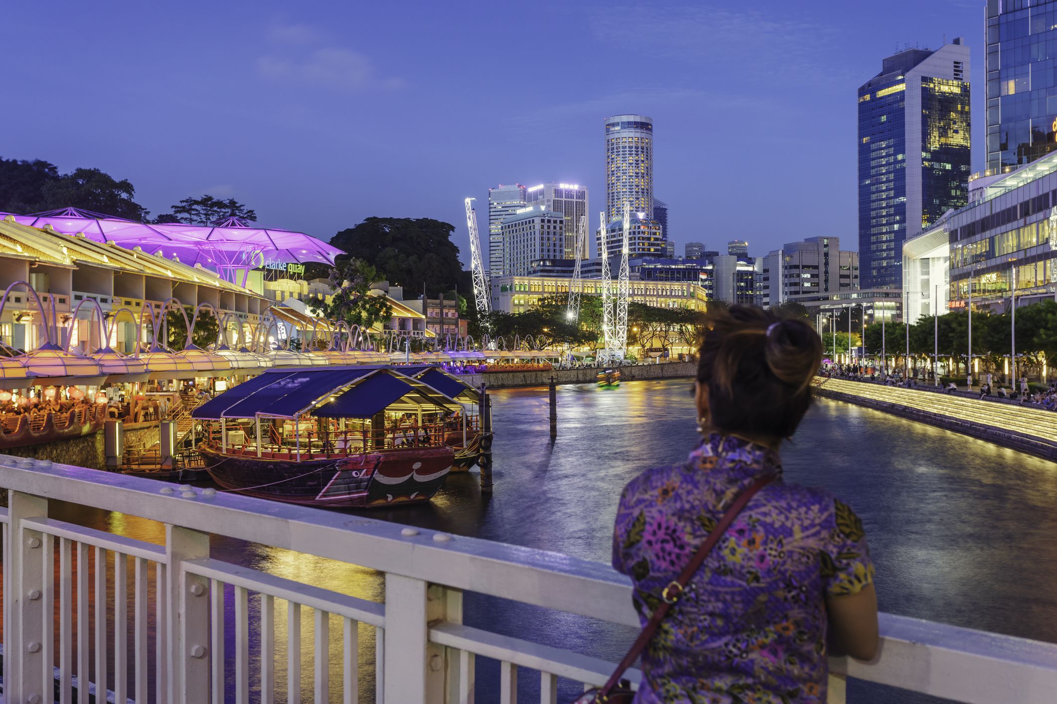 A woman stands by the railing of Read Bridge looking towards Clarke Quay, a historical quay popular for its nightlife with many restaurants and pubs.