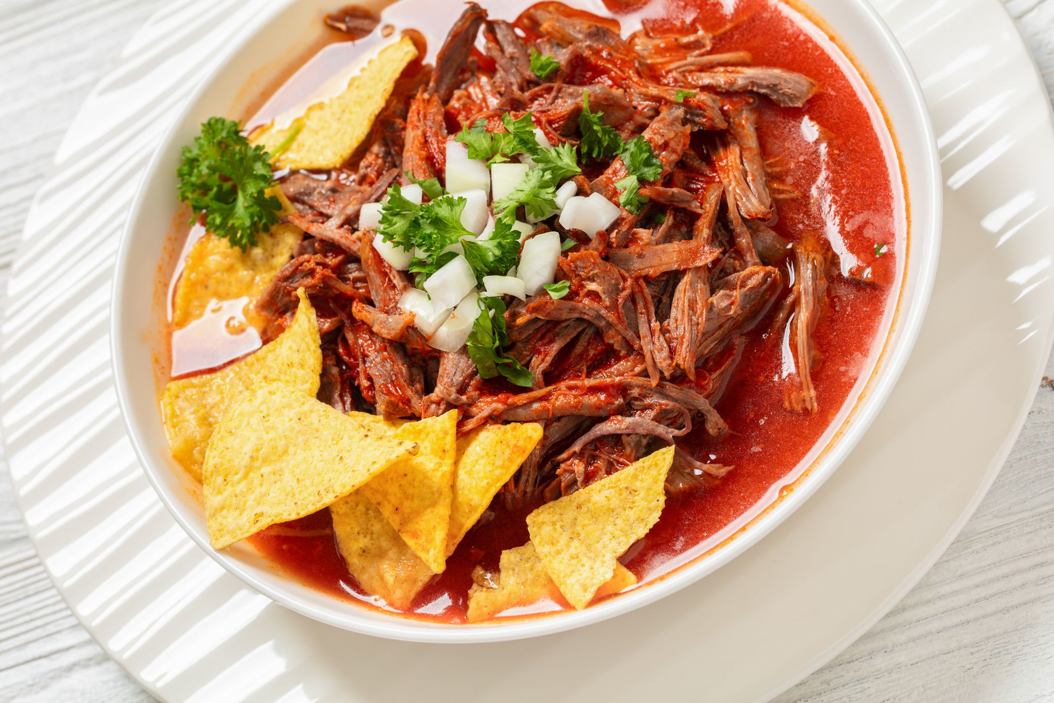 birria de res, mexican beef stew in hot red pepper sauce with raw onion and chopped cilantro, taco chips in white bowl, close-up, dutch angle vew