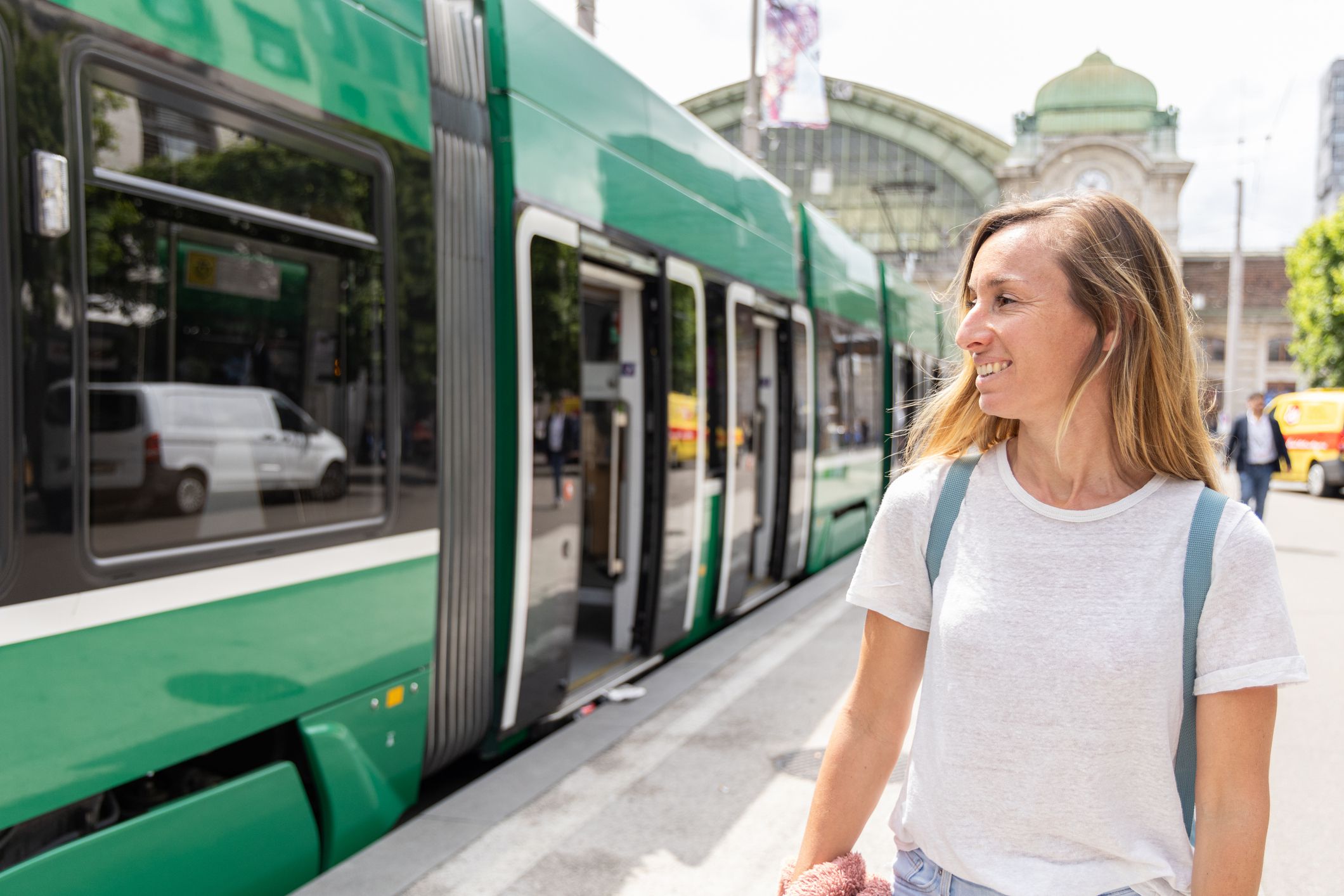 A woman in a white shirt and backpack smiles as she stands next to a green train at a bustling train station on a sunny day.