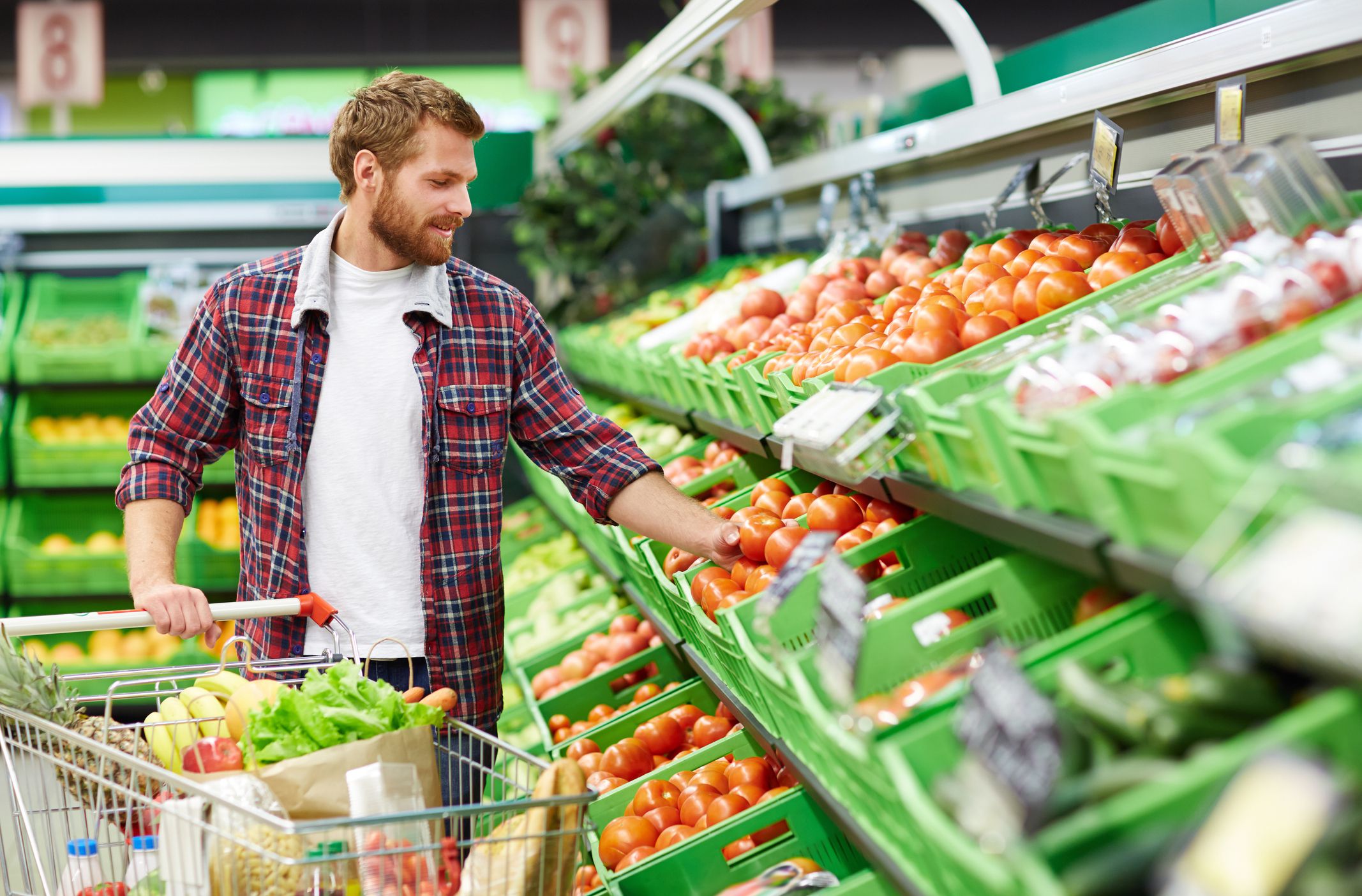 Handsome man with shopping trolley touching tomato in order to define its quality in fruit and vegetable department of supermarket