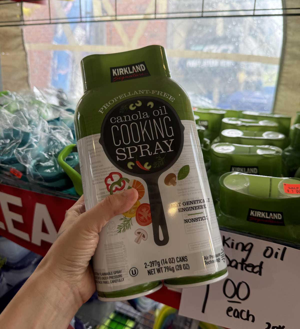 A hand holds a Kirkland canola oil cooking spray twin pack in a store, with more cooking spray packs on the shelf and a handwritten price sign in the background—one of the Costco products to avoid, according to some savvy shoppers.