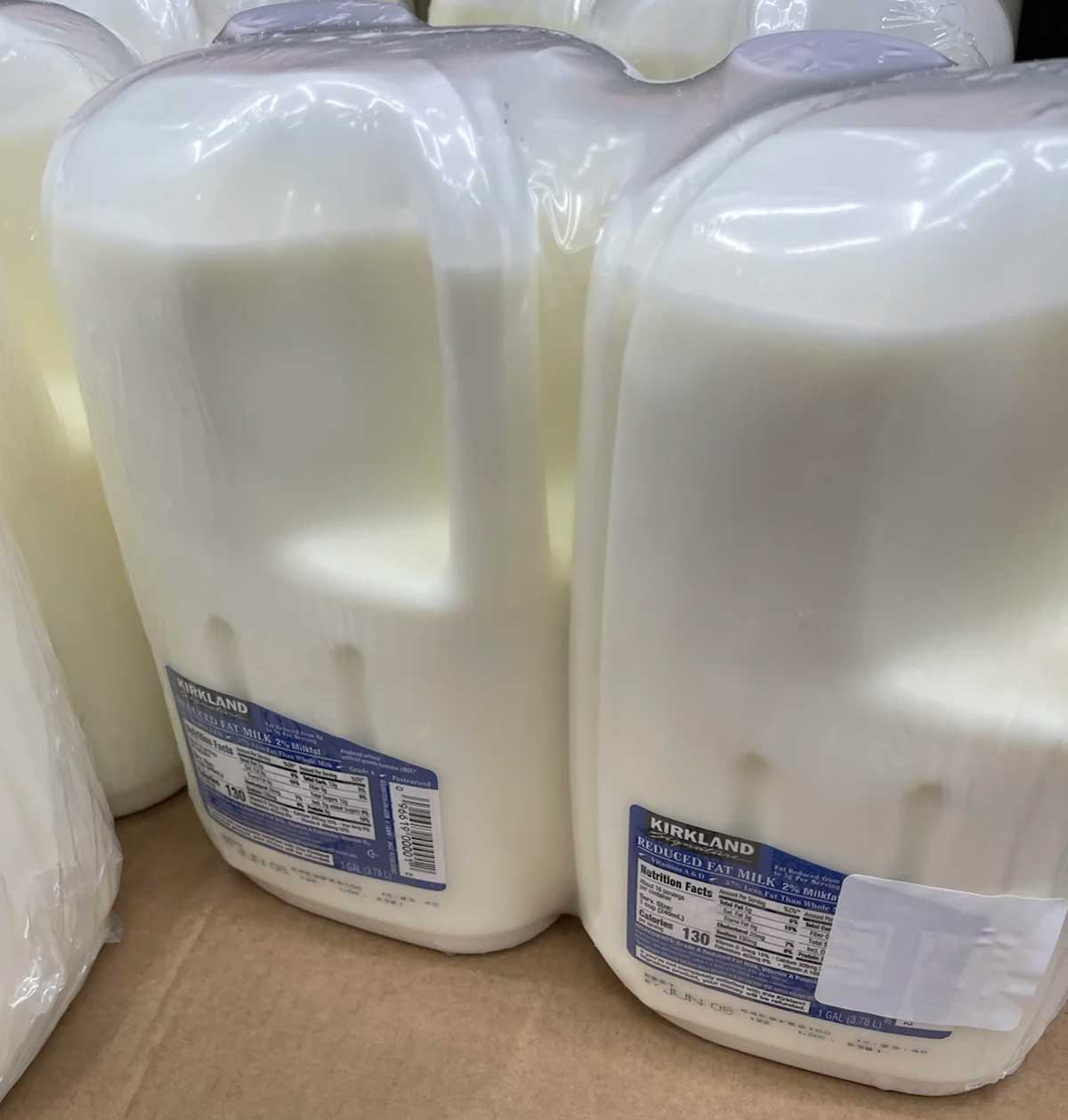 Two large plastic jugs of Kirkland brand whole milk, often mentioned in Costco products to avoid lists, are packaged together on a cardboard surface. The nutrition labels and blue branding are clearly visible on the front of each jug.