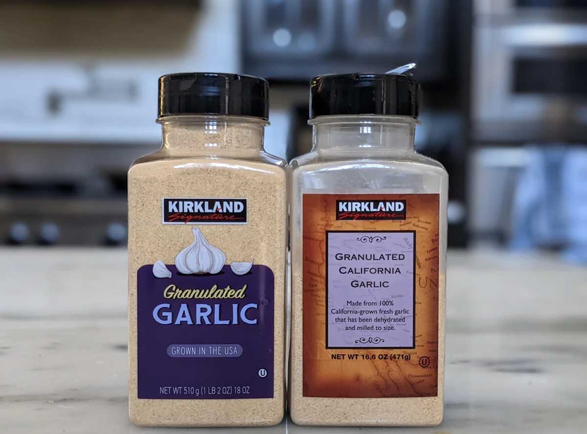 Two large plastic containers of Kirkland brand granulated garlic, often mentioned among Costco products to avoid, sit side by side on a kitchen counter—one with a purple label and one with a brown label. A stove and appliances are blurred in the background.