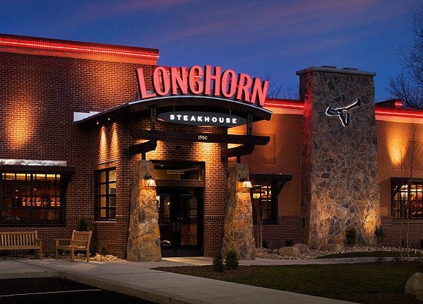 Exterior of a LongHorn Steakhouse restaurant at dusk, featuring illuminated signage, stone columns at the entrance, and warm lighting from windows and a neon longhorn logo on the building.