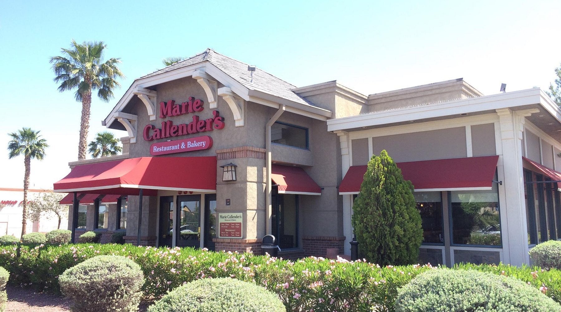 Marie Callender’s Restaurant & Bakery building with red awnings, large front sign, landscaped bushes, and palm trees under a clear blue sky.