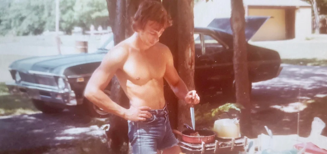 A shirtless man wearing denim shorts cooks at an outdoor stove in a sunny area, with a black car parked nearby and trees providing shade.