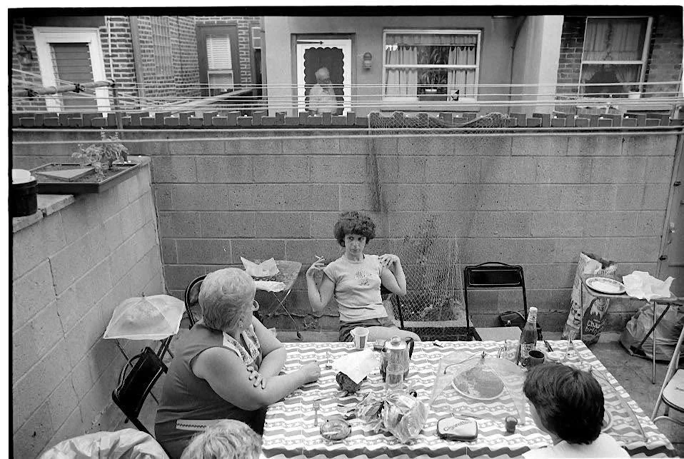 Four people sit around a table with a checkered tablecloth in a small backyard patio, surrounded by cinderblock walls, food, and drinks. One person sits back alone, gesturing, while three others face her.