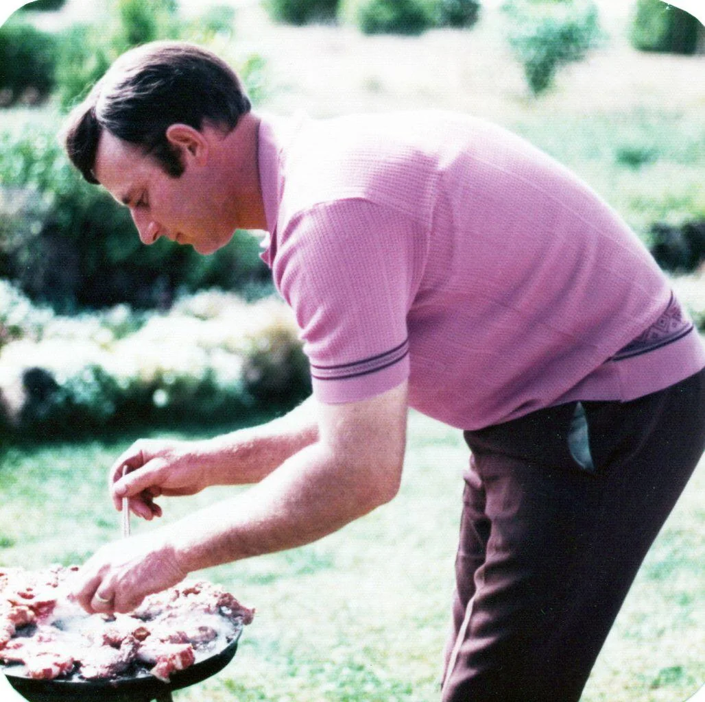 A man in a short-sleeve pink polo shirt and brown pants is preparing food on a round outdoor grill in a sunny garden setting.