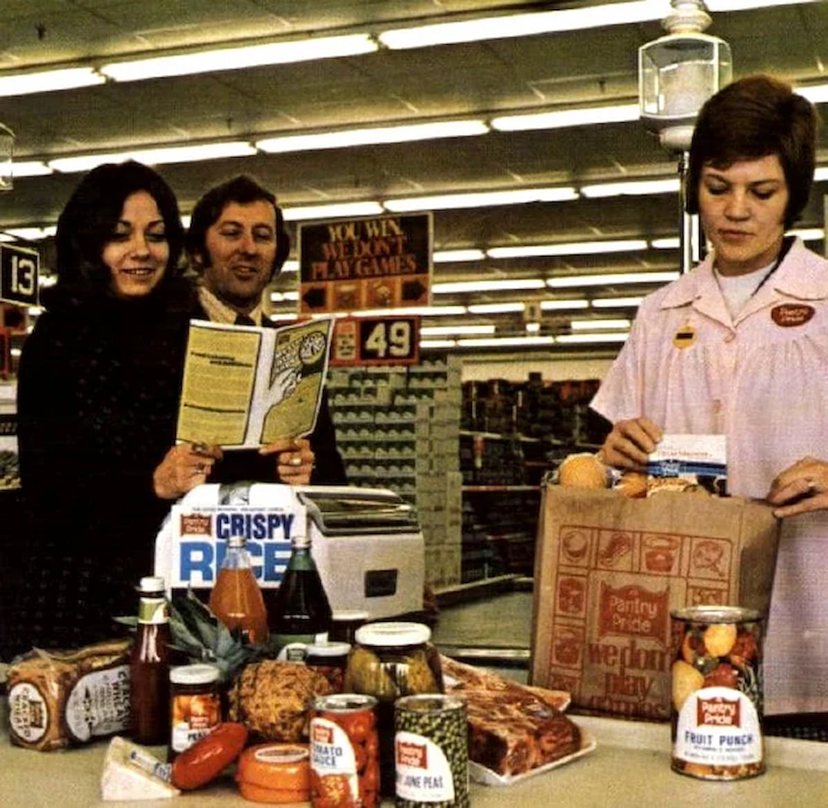 A woman and man stand at a grocery store checkout, smiling and holding a pamphlet, glad to shop local instead of facing closed grocery stores. A cashier in a pink uniform bags rice, ketchup, and vegetables on the counter.