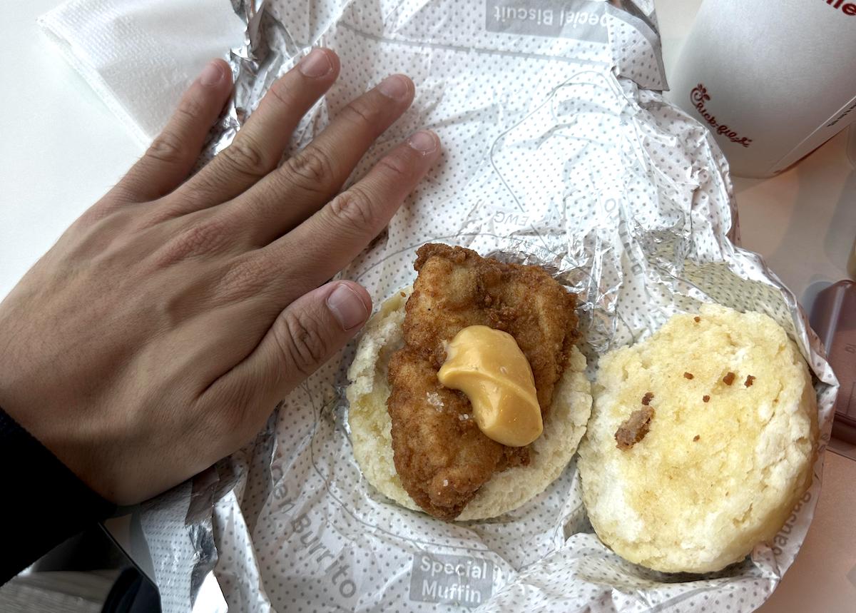 A hand next to an open biscuit sandwich on wrapper paper, showing a piece of fried chicken—like those from some of the worst chicken chains—and a slice of cheese on one half, with the other half of the biscuit beside it. A drink cup is partially visible in the background.