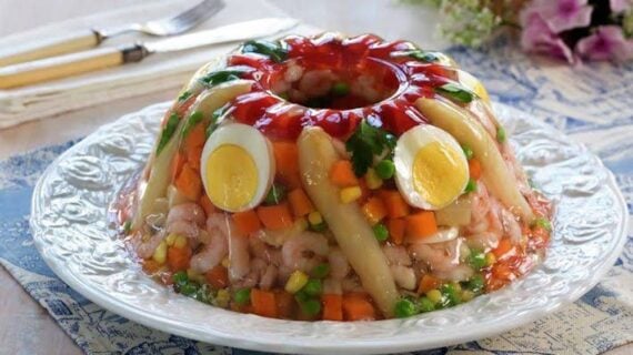A colorful gelatin salad mold with shrimp, vegetables, hard-boiled eggs, and asparagus on a white plate, placed on a blue and white tablecloth next to cutlery and flowers.