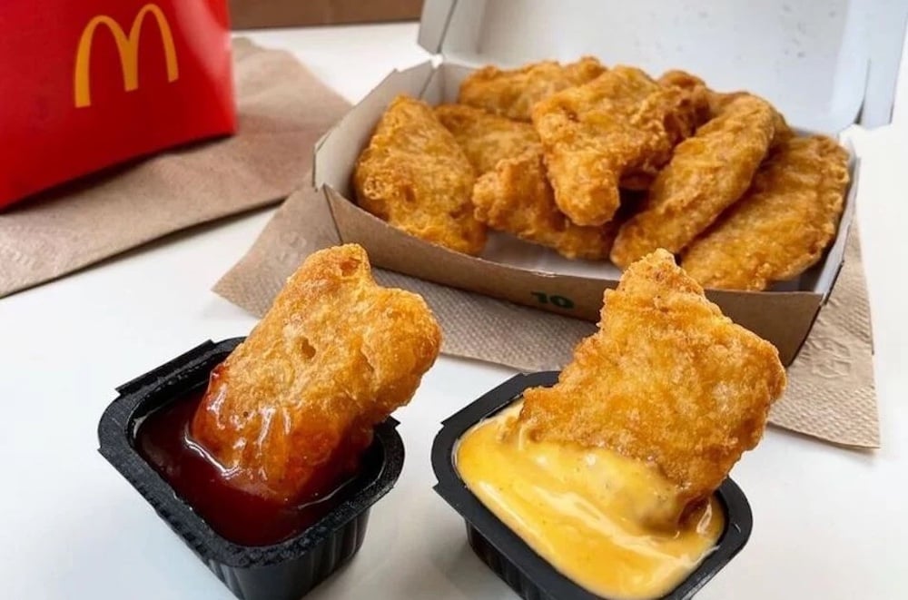 A box of the best fast-food chicken nuggets from McDonald's, with two nuggets being dipped—one in barbecue sauce and the other in cheese sauce—sits on a table beside a red McDonald's fries container.