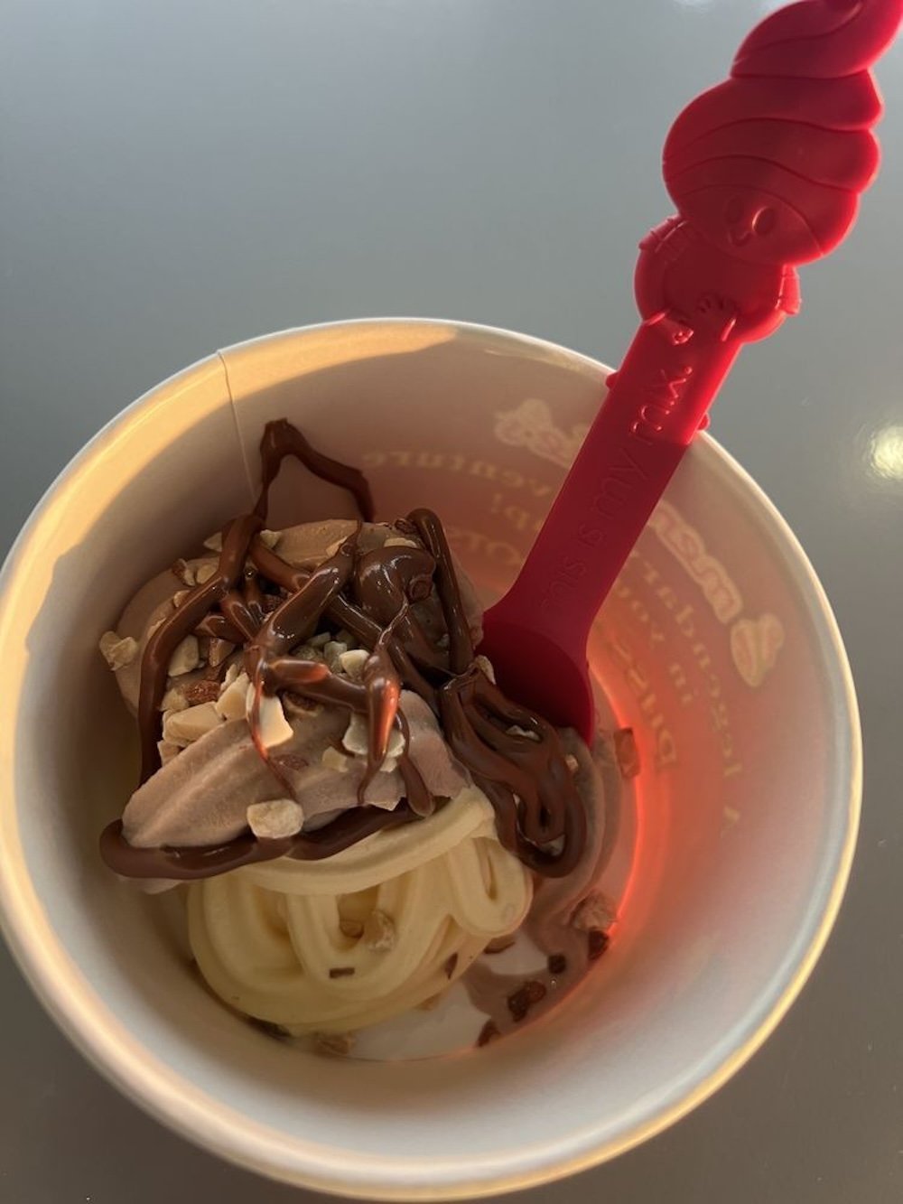 A cup of soft serve ice cream topped with chocolate sauce and chopped nuts, served with a red spoon shaped like an ice cream swirl.