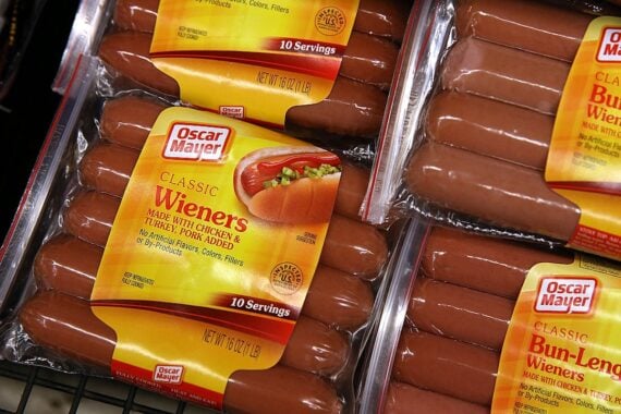 Packages of Oscar Mayer Classic Wieners are displayed on a store shelf. The packaging is yellow and clear, showing the hot dogs inside, and includes serving size and ingredient information.