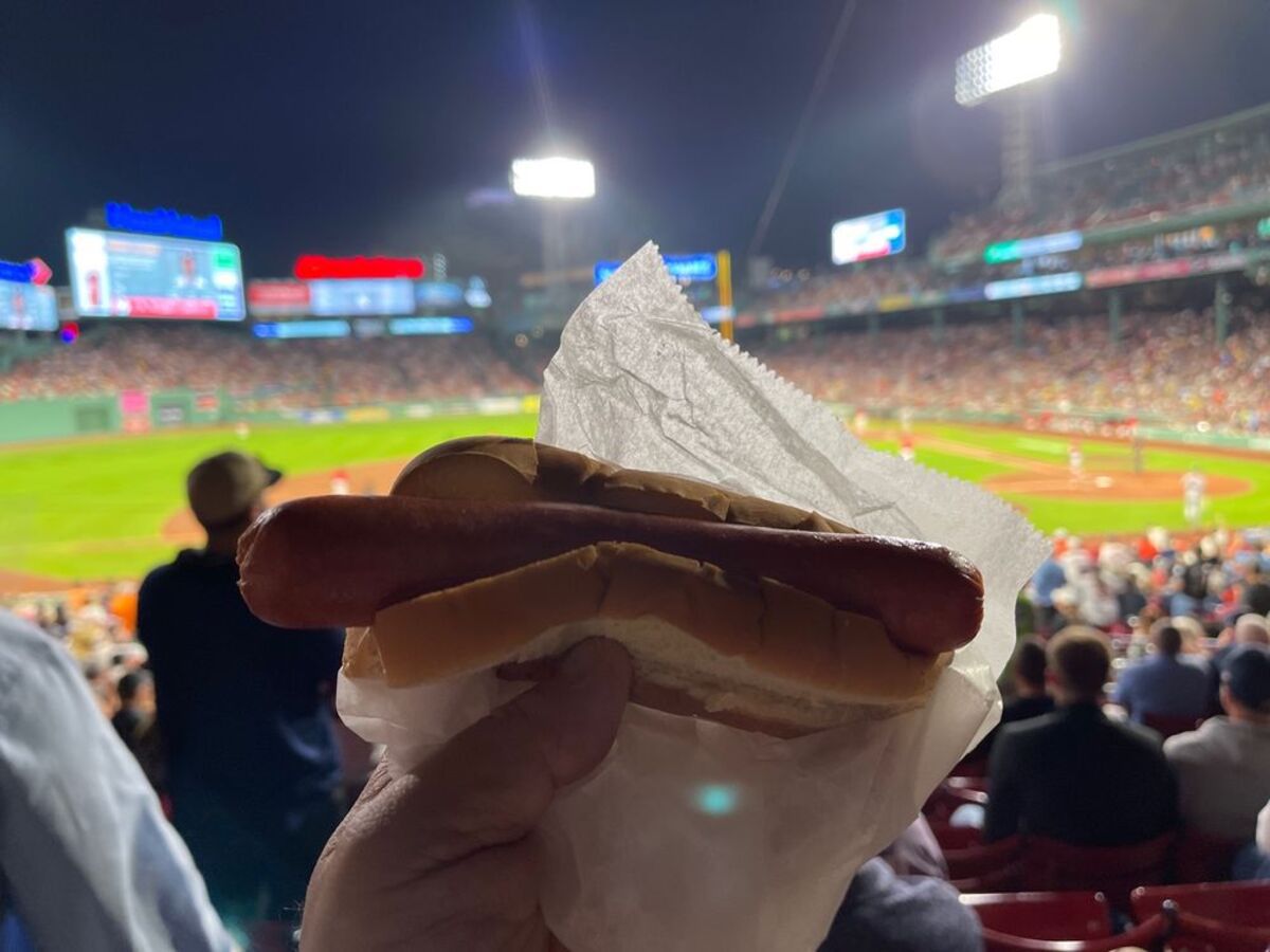 Hand holding a Fenway Frank in white serving paper, in the stands during a baseball game at Fenway Park, Boston, during night, field is lit in the background