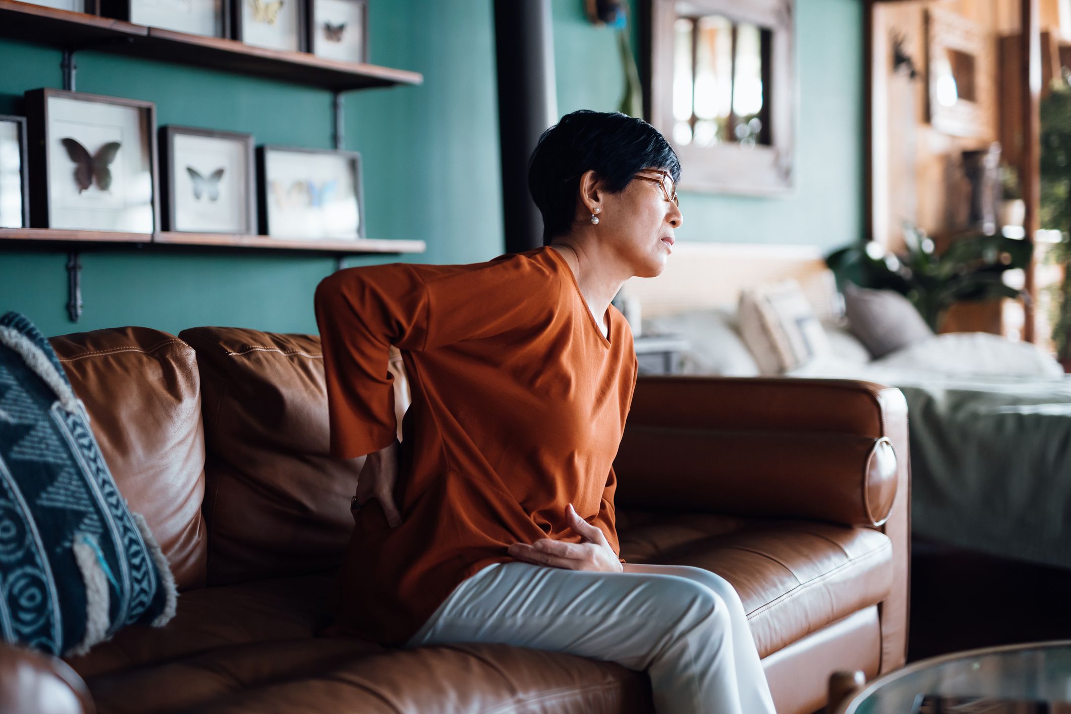 A distraught senior Asian woman feeling unwell, suffering from backache, massaging aching muscles while sitting on sofa in the living room at home. Elderly and health issues concept