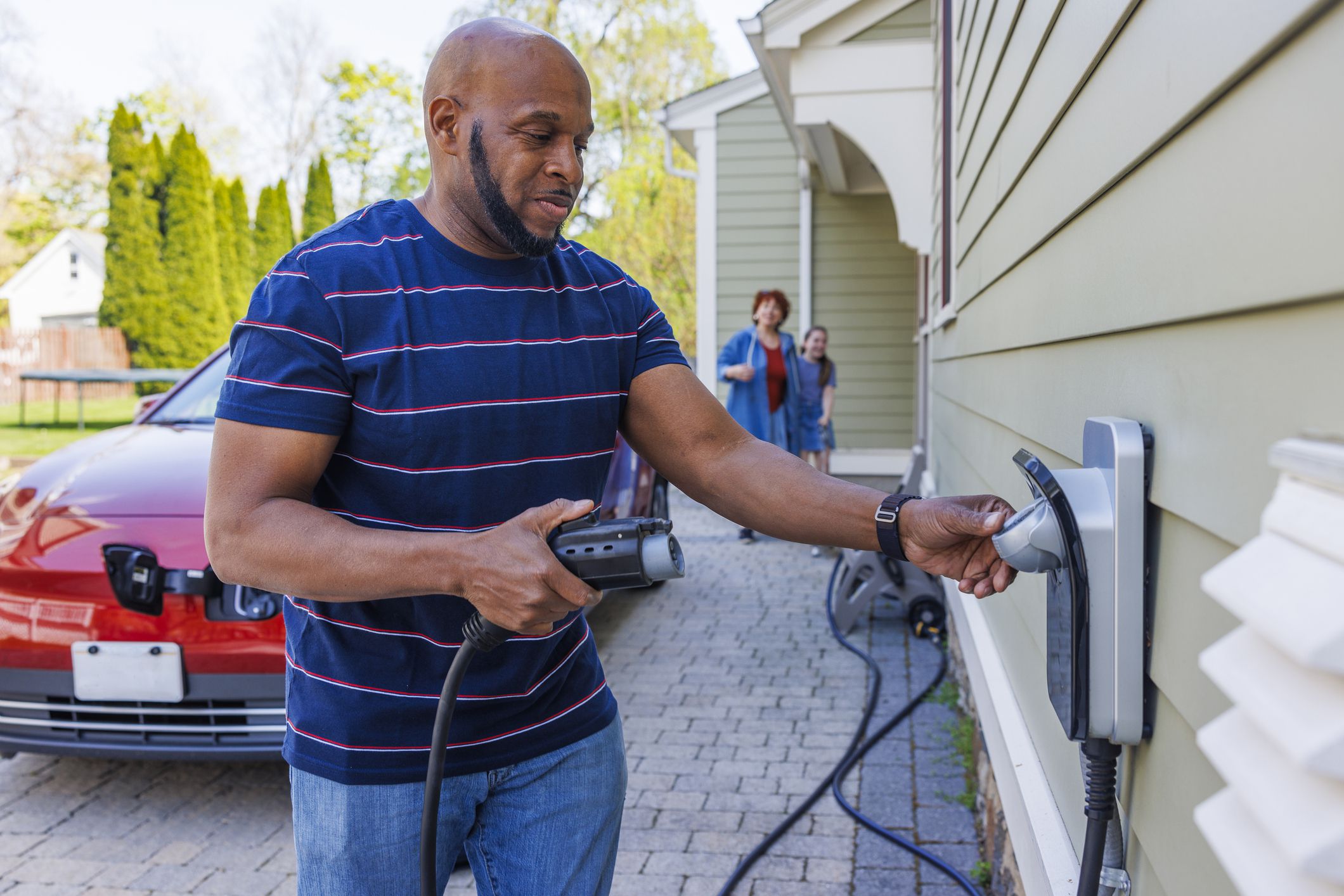 A middle-aged African-American man smiling as he plugs an electric charger and prepares the power cable in the driveway of a suburban home in Madison, New Jersey. The background shows his interracial family walking around and approaching