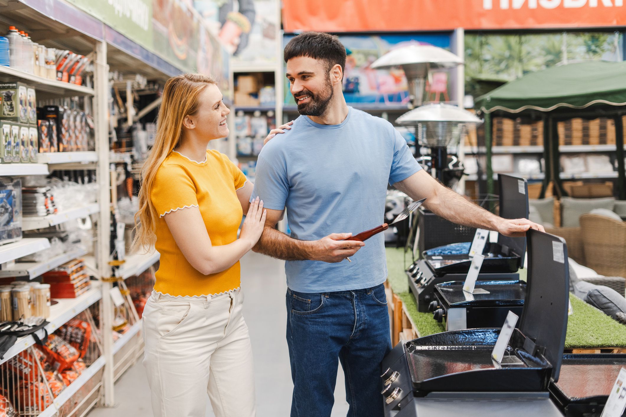 Portrait of smiling young man and woman choosing barbeque at electric store while looking each other, buying barbecue appliance
