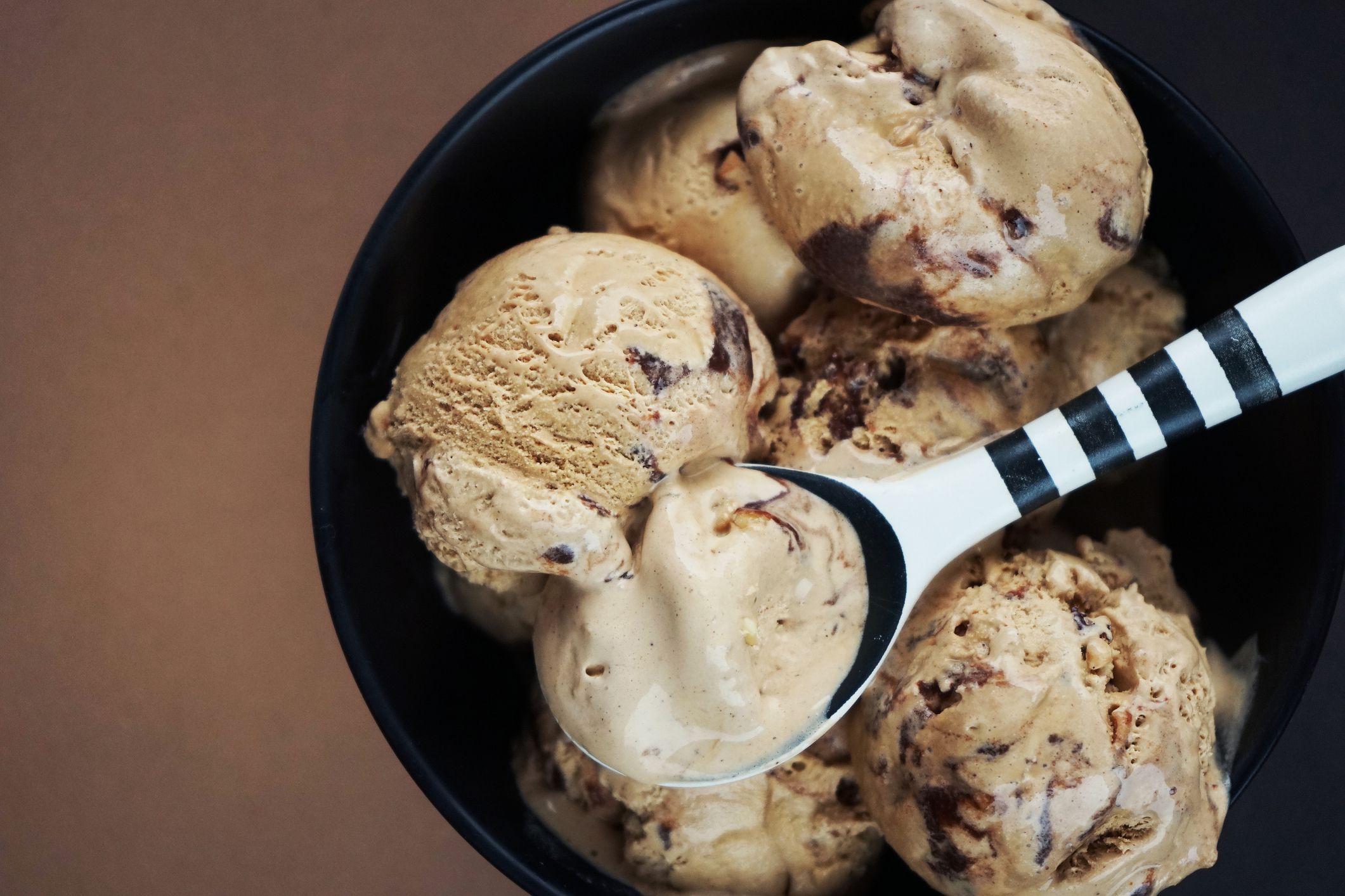 Nut ice cream balls with chocolate in a black bowl and a black and white spoon on a brown background