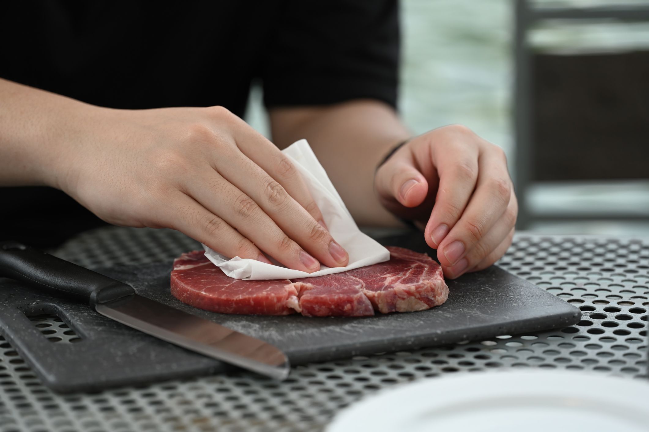 Drying Steak for a Perfect Cook, Hands gently patting down a raw steak to dry off with a paper towel on a cutting board.