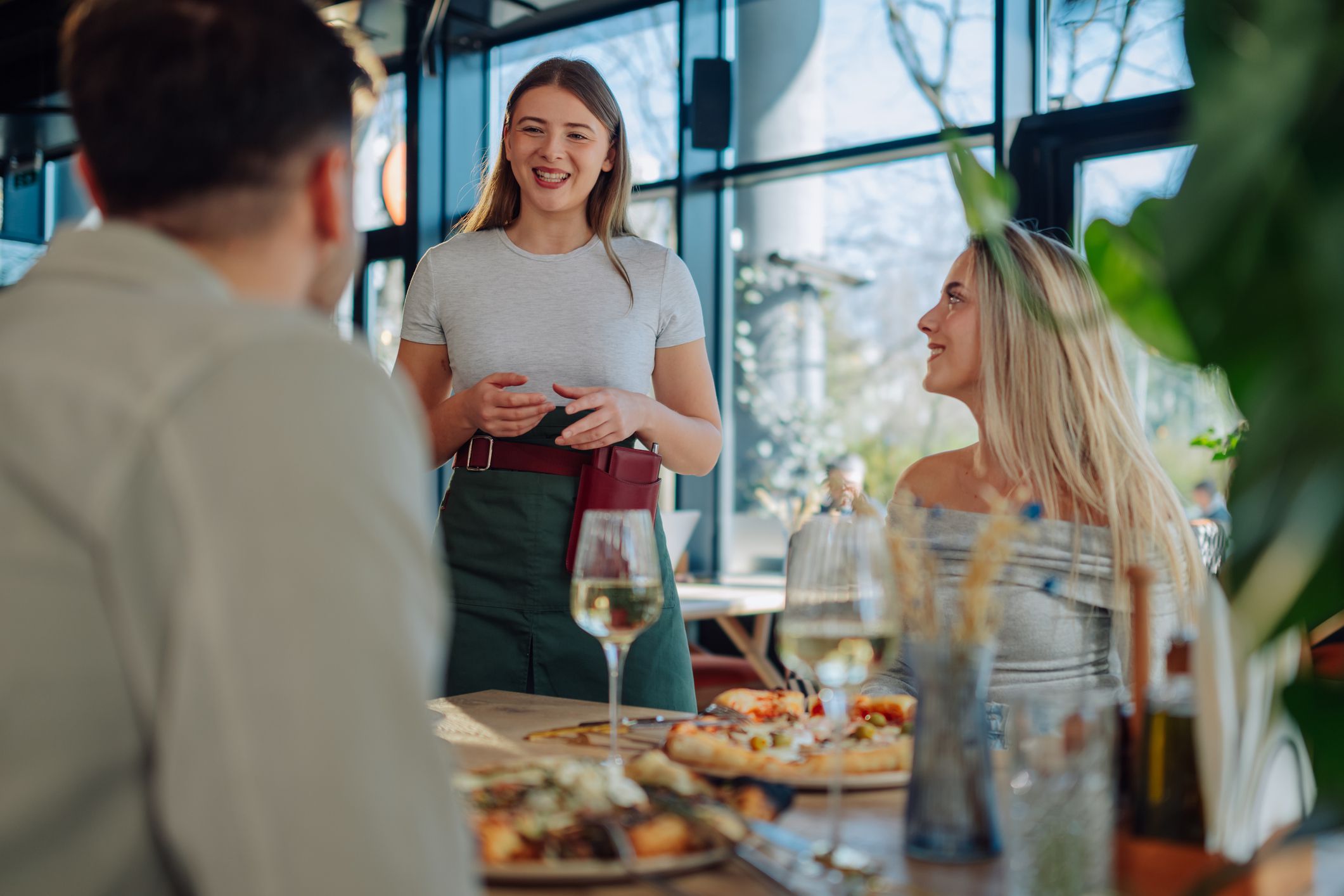 Young waitress taking order from customers sitting at a table, enjoying pizza and wine in a modern restaurant