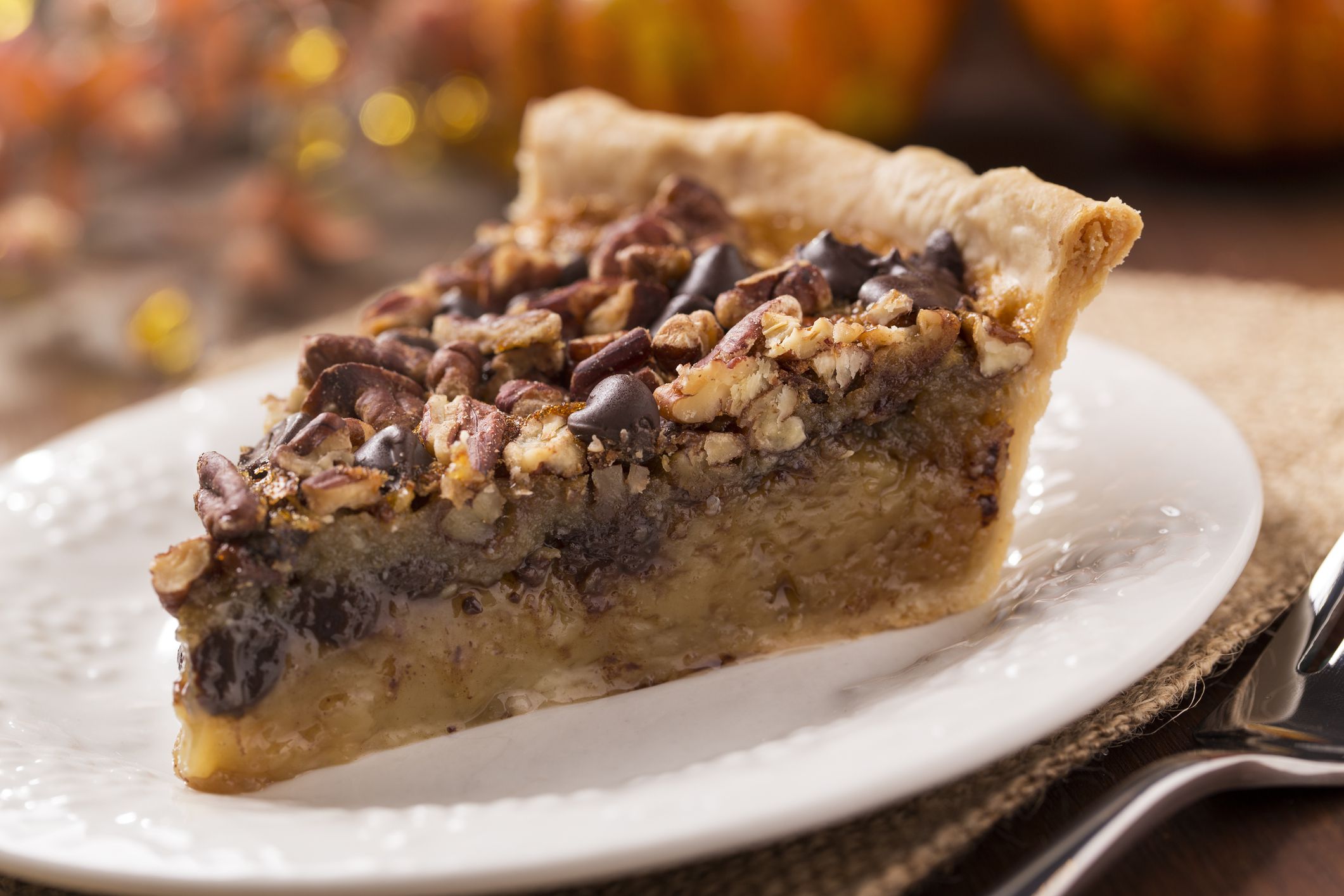 A slice of chocolate pecan pie on a plate with a burlap place setting and fork. In the background is Thanksgiving / Fall decorations. The decorations are a variety of pumpkins and gourds. The shot is horizontal with selective focus.