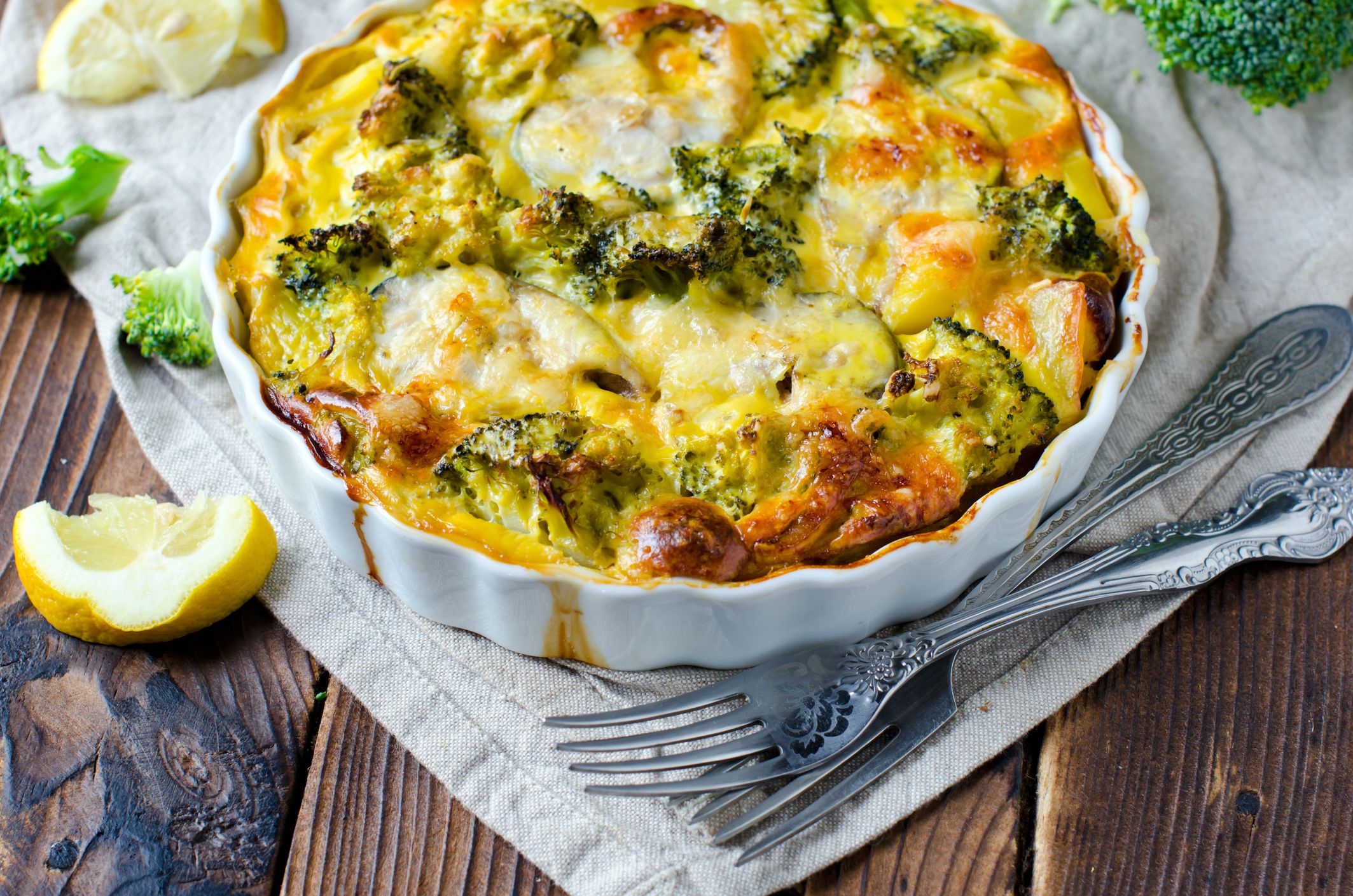A colorful fish and broccoli casserole in a white baking dish. The casserole sits on a plank style wooden surface with lemon wedges and forks.
