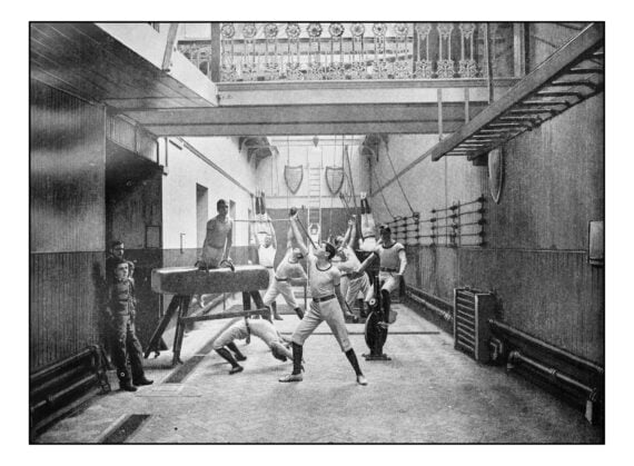 A group of men in a gymnasium practicing various calisthenics and exercises on a pommel horse, parallel bars, and a stationary bicycle.