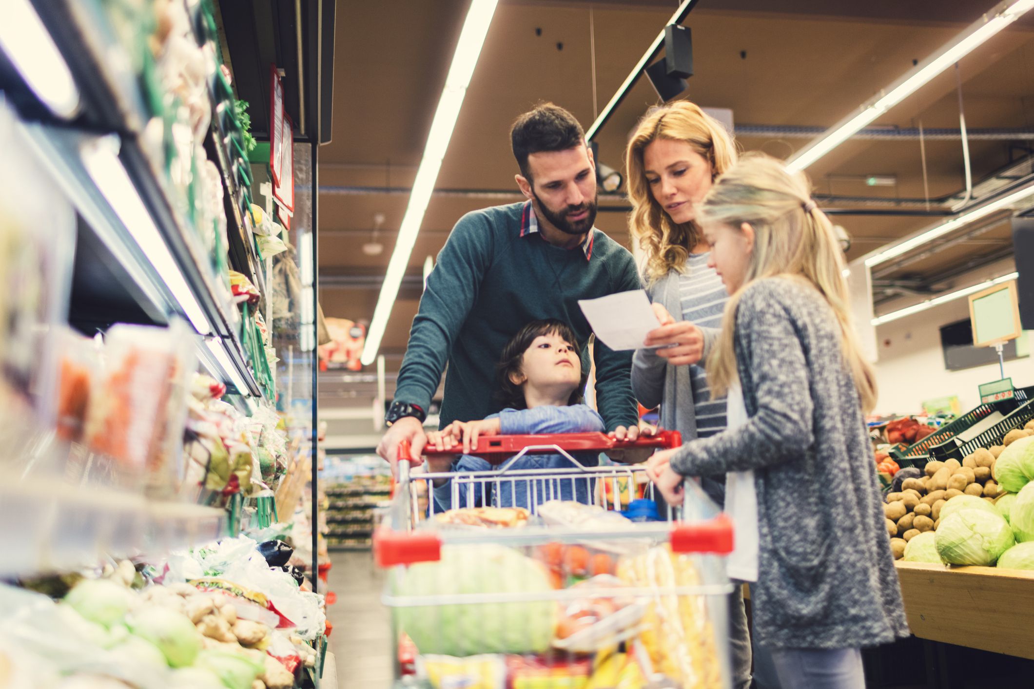 Cheerful family with two children shopping vegetables in supermarket. Holding shopping list and looking for groceries