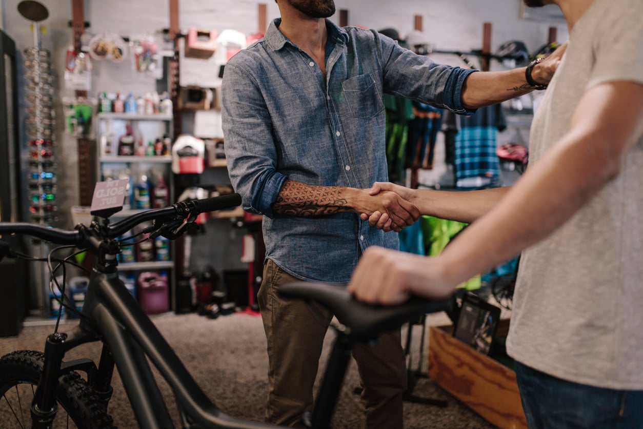 Two people shaking hands in a bike shop, with one holding a bicycle. Shelves with cycling gear and accessories are visible in the background.