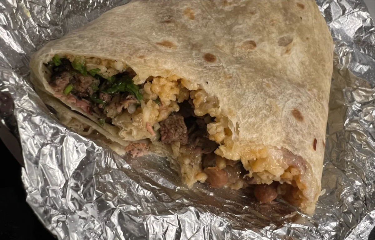 A partially eaten burrito rests on crinkled foil, revealing fillings of rice, beans, grilled meat, and greens wrapped in a flour tortilla.