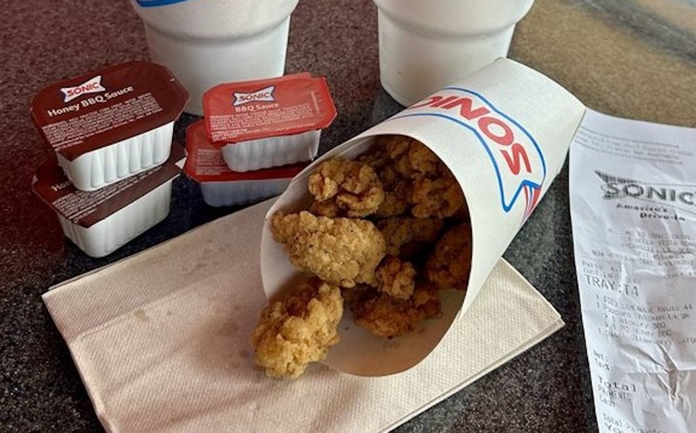 A container of fried chicken pieces from Sonic, often ranked among the best fast-food chicken nuggets, sits on a napkin next to a receipt, three dipping sauce containers, and two white drink cups with red straws.