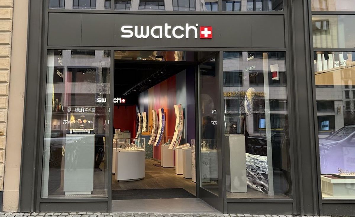 Street view of a Swatch store with large glass doors open, displaying watch displays and colorful interior decor inside. The Swatch logo with a red and white Swiss flag is above the entrance.