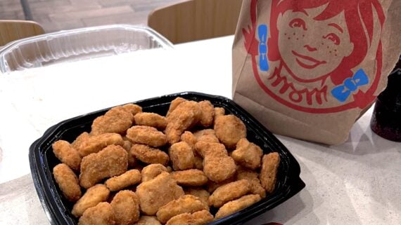 A black tray filled with many of the best fast-food chicken nuggets sits on a table next to a Wendy's brown paper bag with the restaurant's red logo. Part of a plastic container and drink cup are also visible in the background.