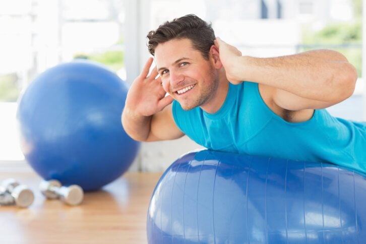 side view portrait of a young man exercising on fitness ball at a bright gym