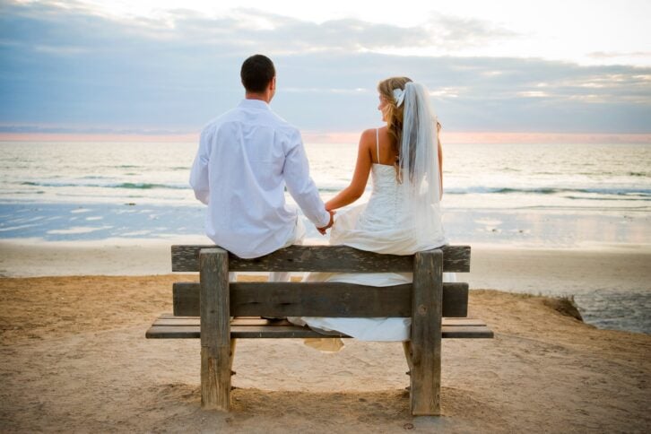 Woman in a wedding dress with man sitting on a bench on the beach