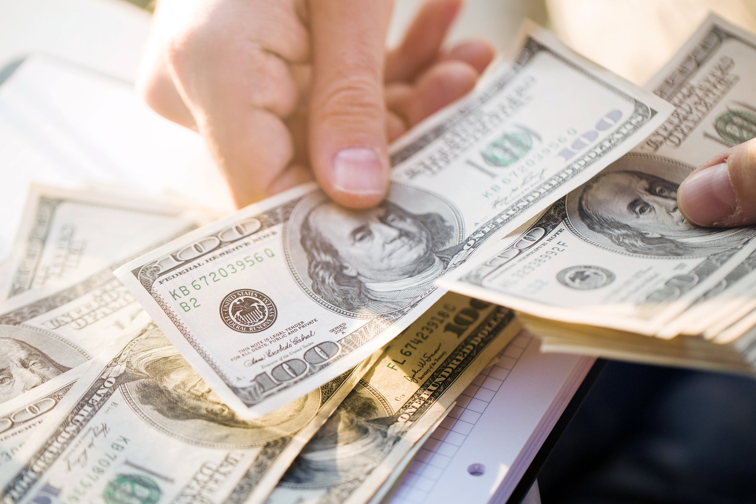 A person’s hands are counting a stack of US $100 bills over a table with more money scattered below. The bills prominently feature Benjamin Franklin’s portrait.