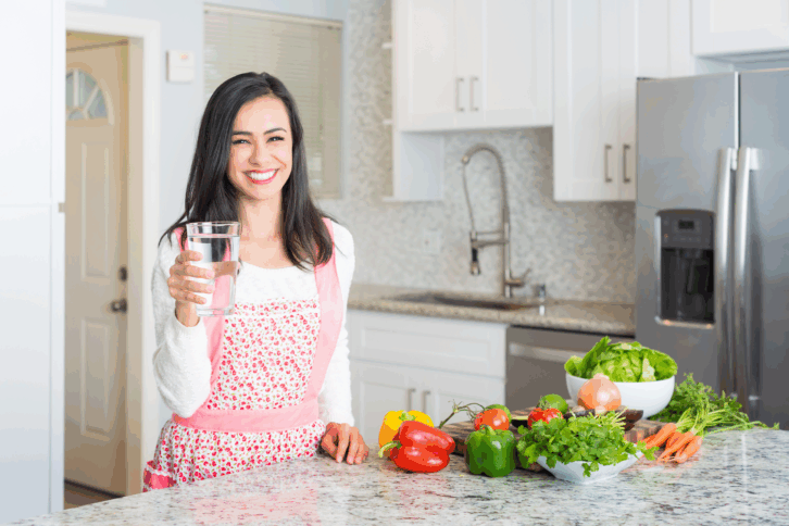 Happy woman in her kitchen holding a glass of water next to an assortment of healthy food