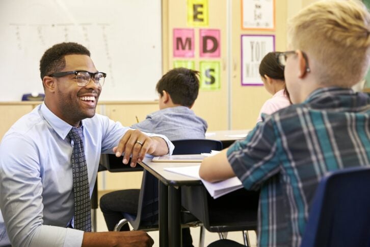 smiling teacher kneeling beside elementary student desk