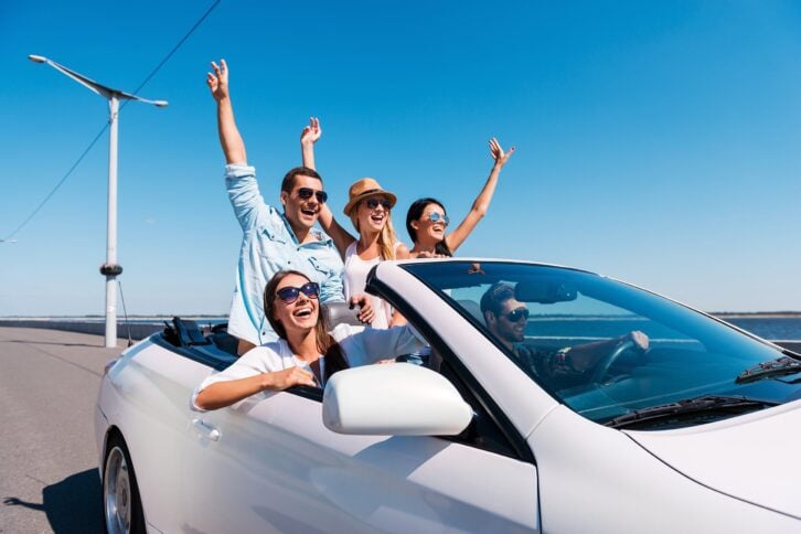group of young happy people enjoying road trip in their white convertible and raising their arms up