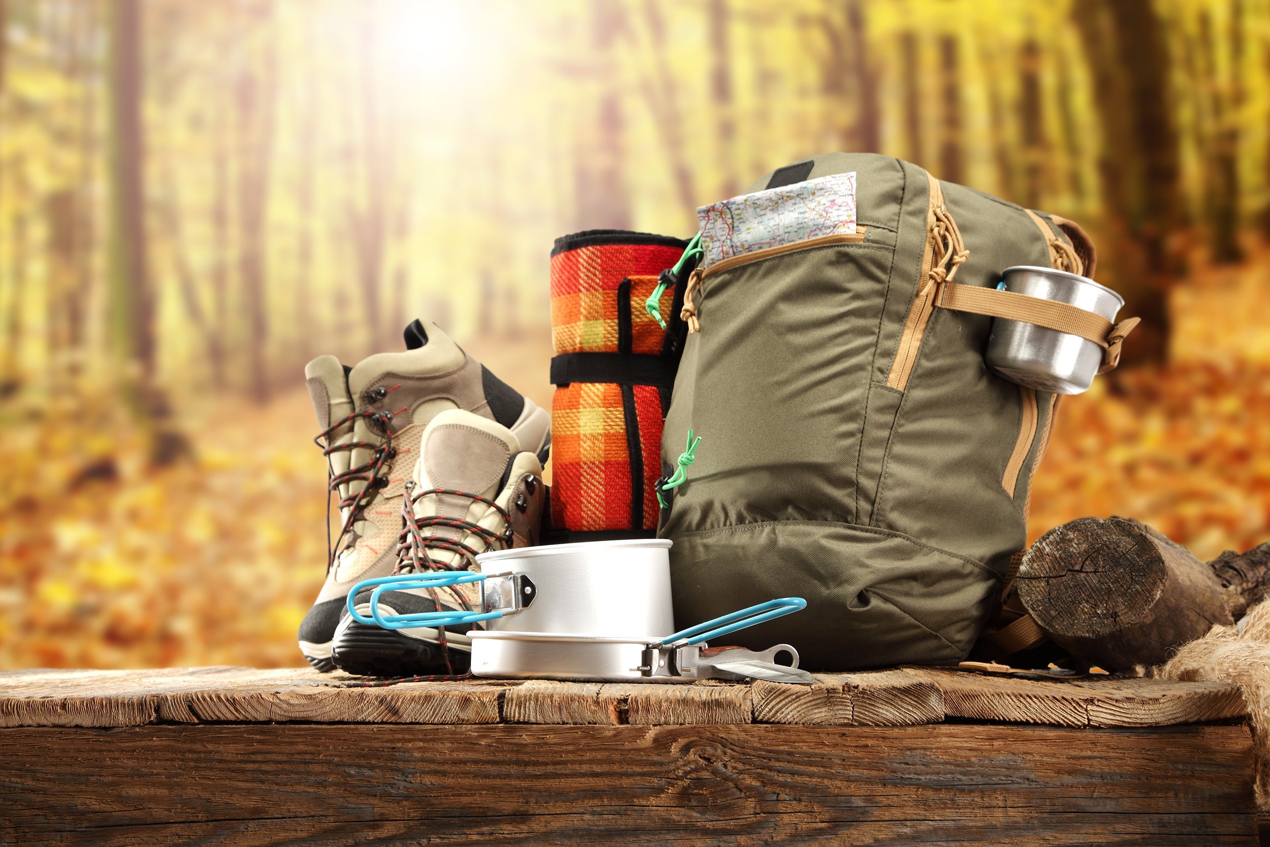 Camping gear including hiking boots, a green backpack with a metal cup, a red plaid blanket, and cooking pots, arranged on a wooden surface with a sunlit autumn forest in the background.