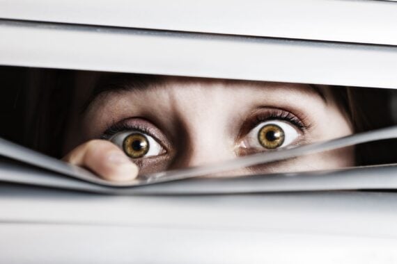 wide-eyed, terrified young woman looks out through venetian blinds