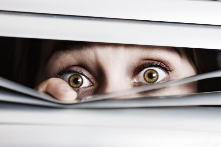 wide-eyed, terrified young woman looks out through venetian blinds