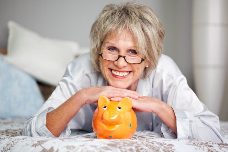 Happy senior female lying on bed with piggybank