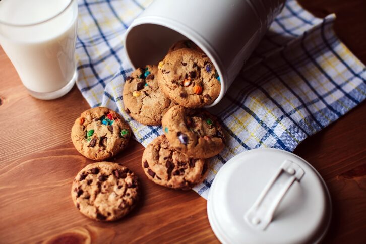 Assorted cookies falling out of a jar on a wooden table with a glass of milk